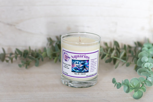 Candle in a glass jar with an Aquarius zodiac label, surrounded by eucalyptus leaves on a light background