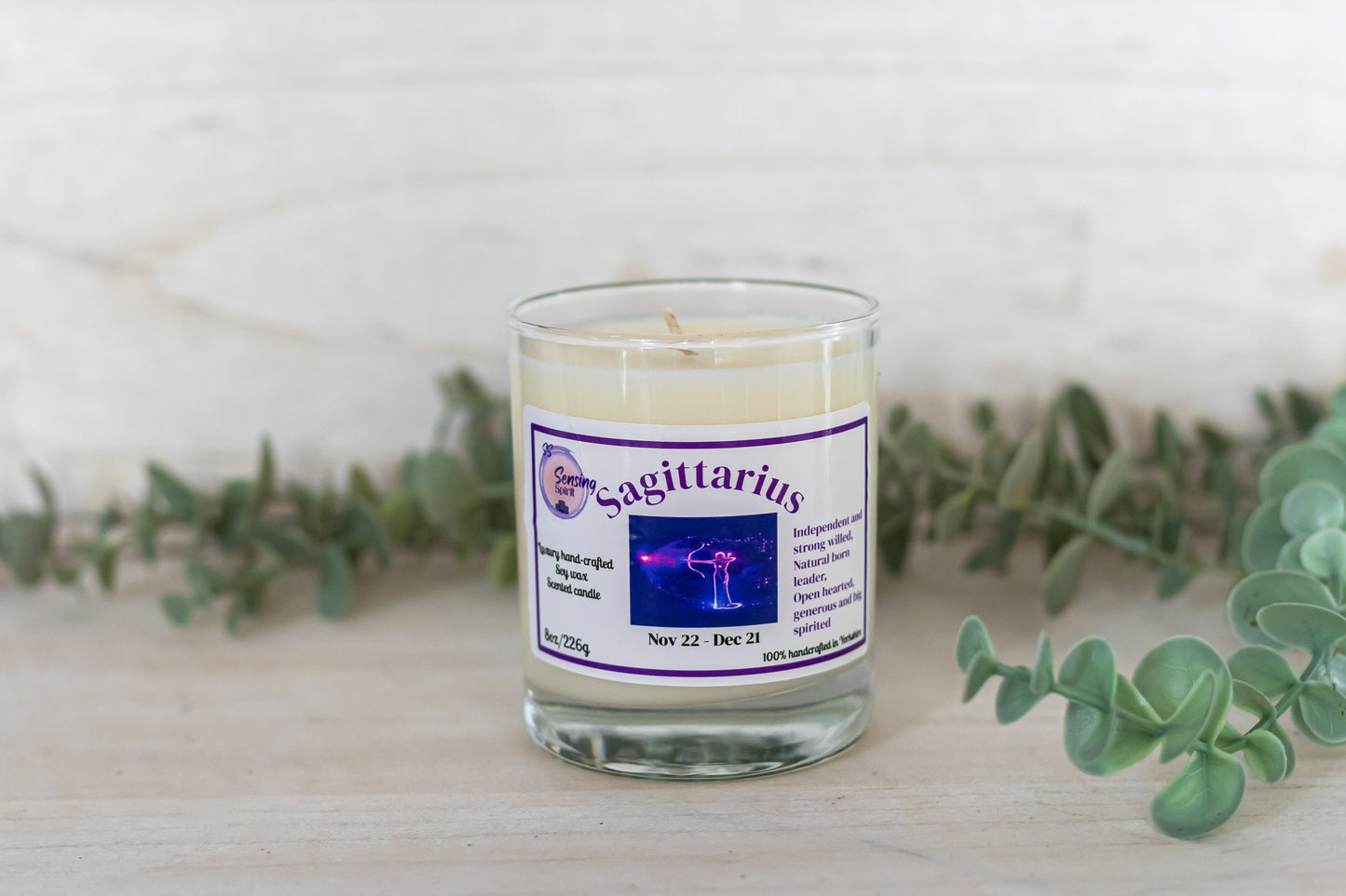 Candle with a 'Sagittarius' label on a white surface with greenery
