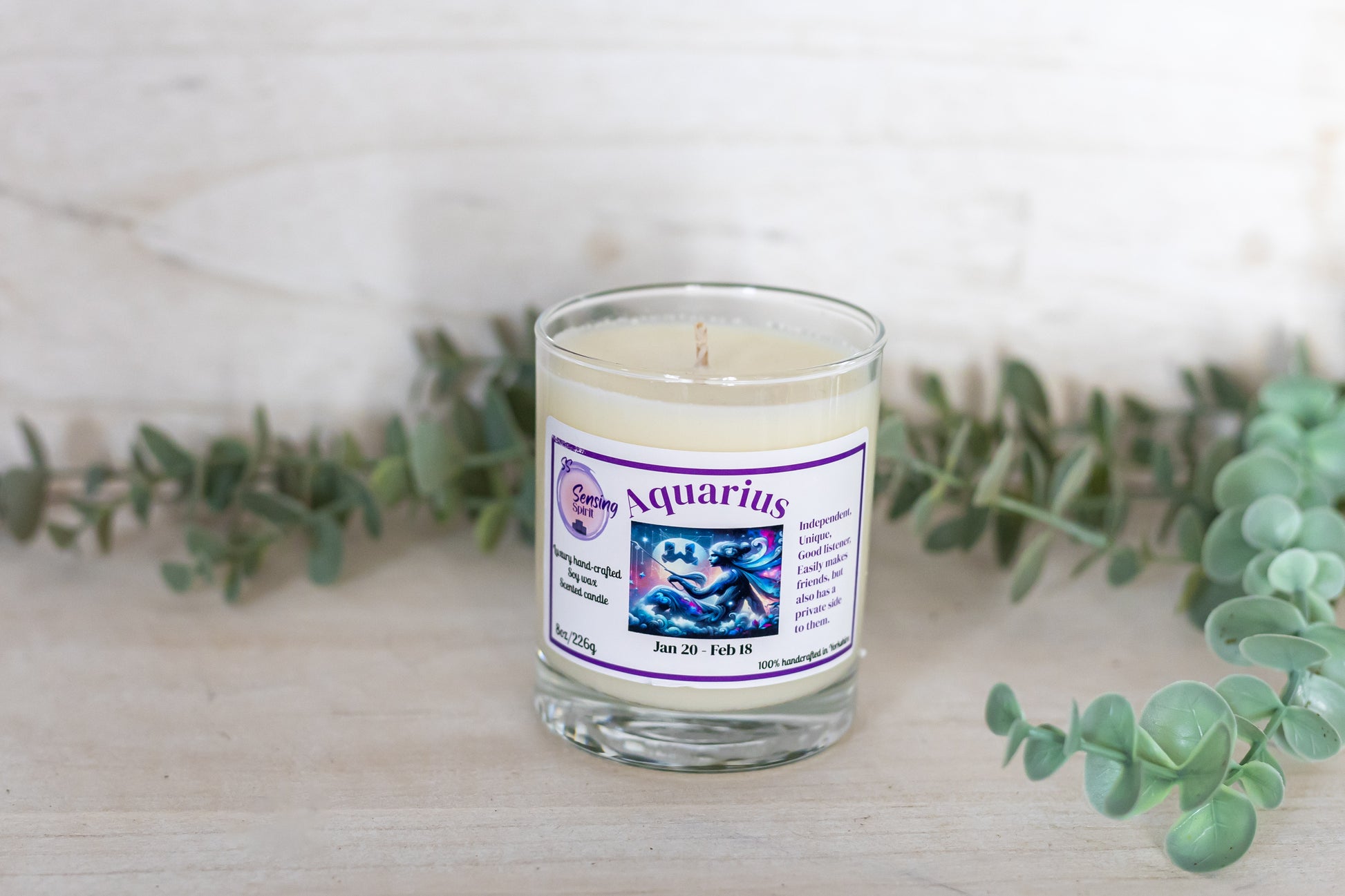 Candle in a glass jar with an Aquarius zodiac label, surrounded by eucalyptus leaves on a light background