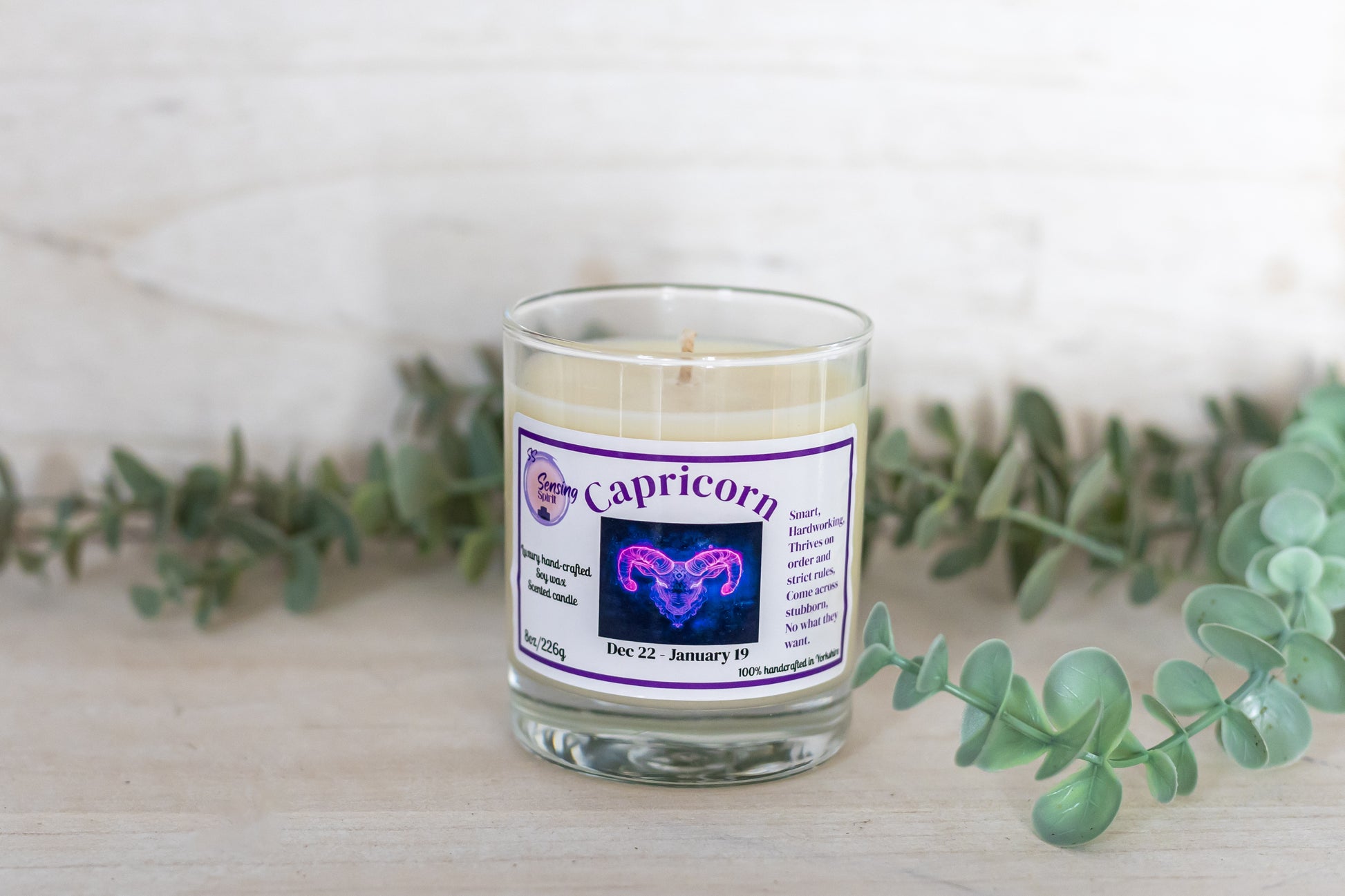 Candle with a Capricorn label on a white surface with greenery