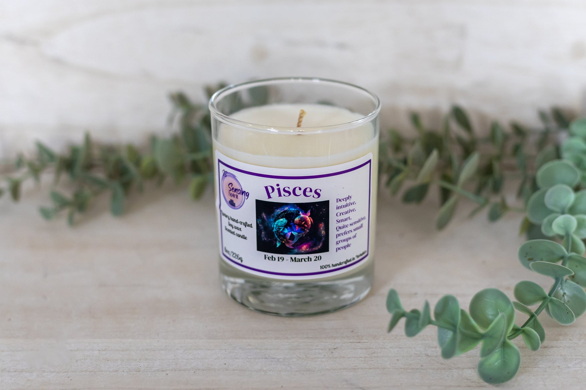 Candle in a glass jar with a 'Pisces' label on a wooden surface with greenery